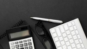 Overhead view of financial tools including a calculator, keyboard, pen, glasses, and notepad on a black surface, symbolizing budgeting or accounting work.
