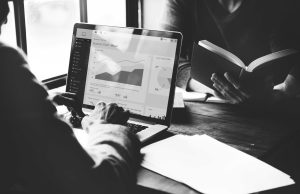 Person analyzing financial charts on a laptop while another person reads a book, indicating investment research or business planning.