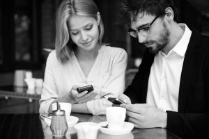 Man and woman sitting at a cafe table using smartphones, possibly managing finances or discussing money-related lifestyle decisions.